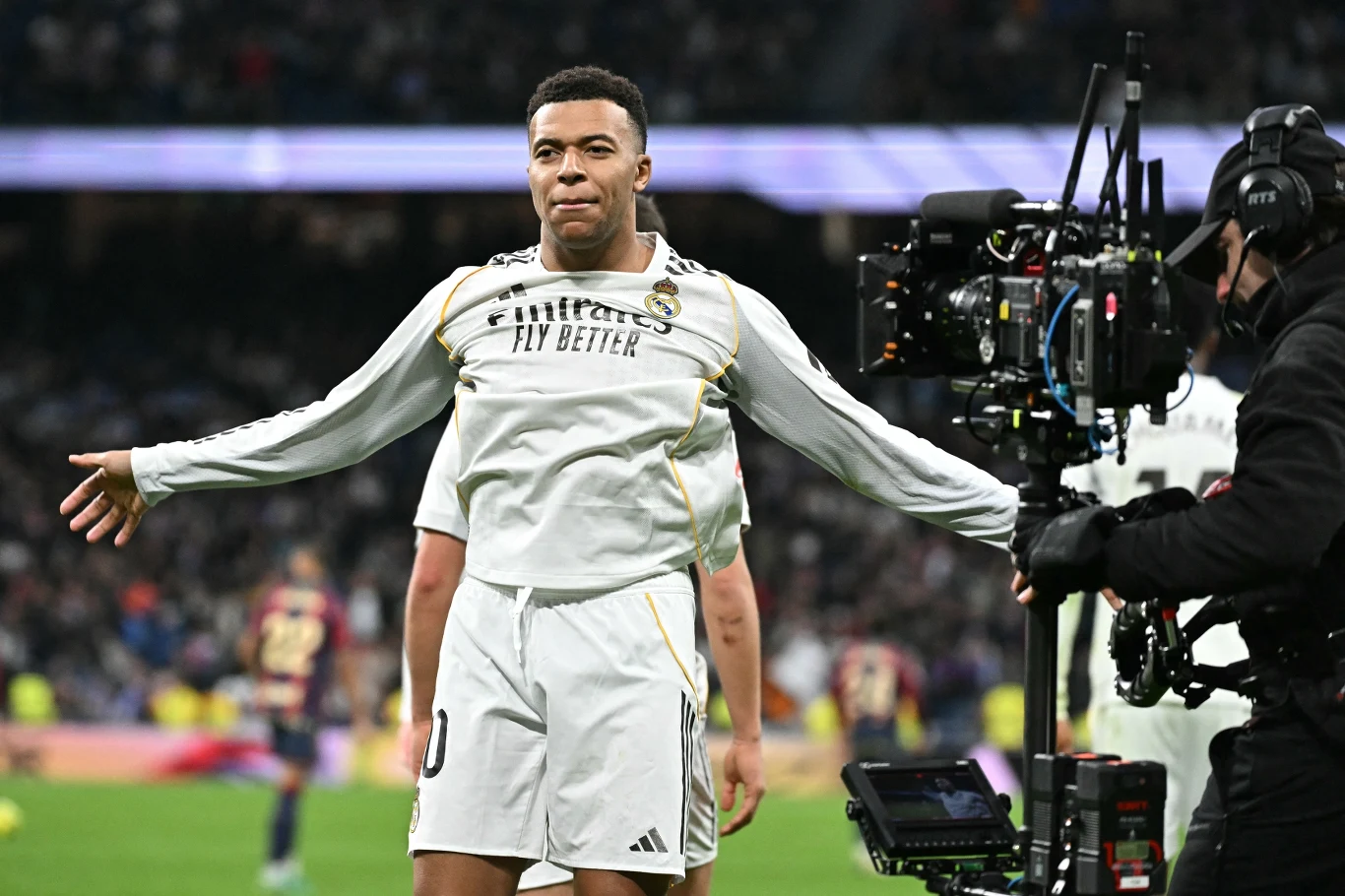 Piłkarz w białym stroju z rozłożonymi rękami świętuje na stadionie podczas meczu piłkarskiego, obok niego widoczna profesjonalna kamera nagrywająca jego zachowanie, w tle rozmazani zawodnicy oraz trybuny pełne kibiców.