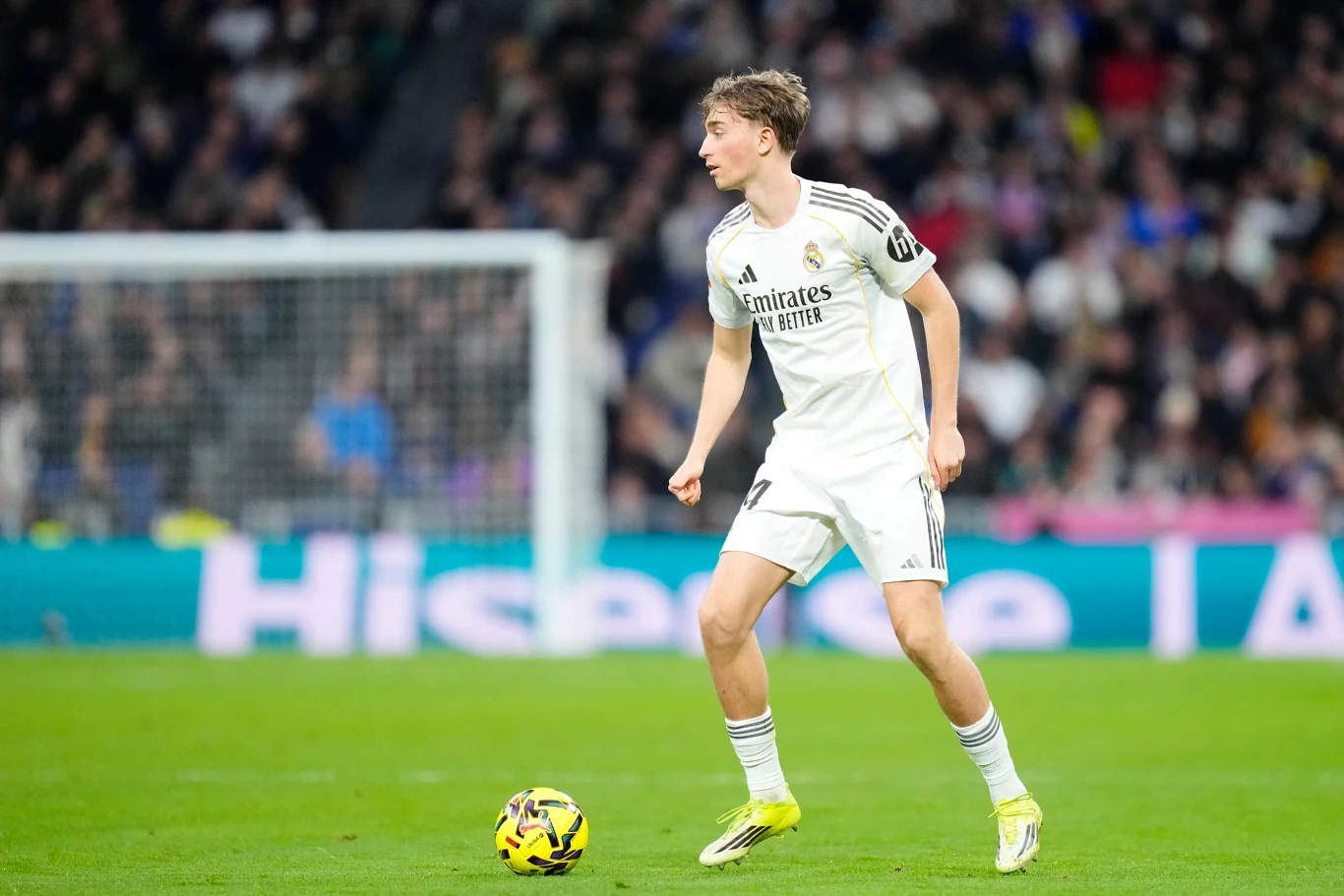 Piłkarz Realu Madryt ubrany w biały strój z logo klubu kontroluje piłkę podczas meczu na stadionie, w tle widoczni kibice na trybunach.