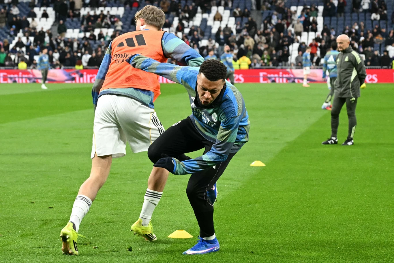 Dwóch piłkarzy wykonuje intensywne ćwiczenia rozgrzewkowe na murawie stadionu, w tle widoczni pozostali członkowie drużyny oraz trybuny z kibicami.