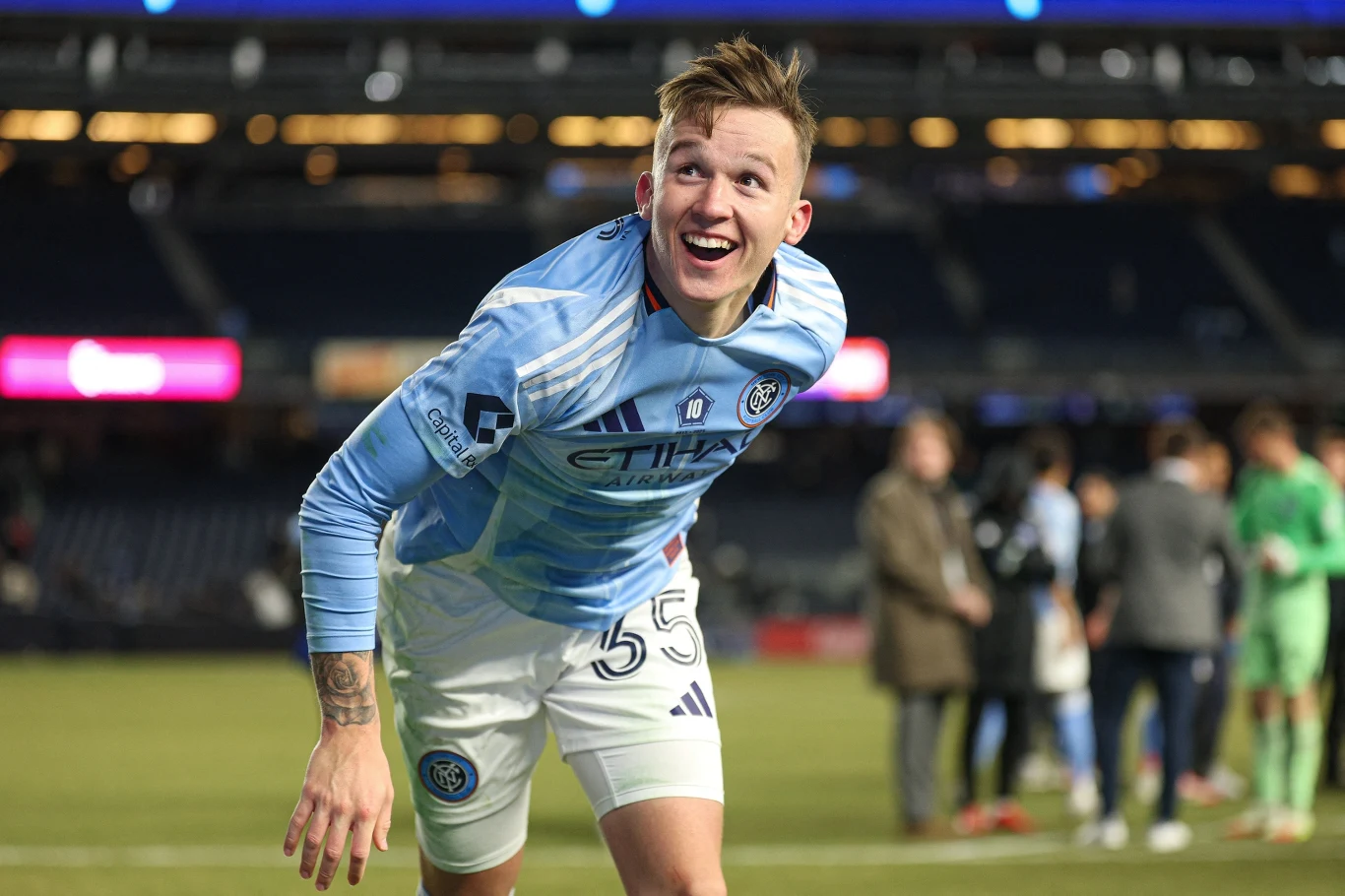 Młody piłkarz w niebieskiej koszulce sportowej numer 55 uśmiecha się szeroko podczas przebywania na stadionie, w tle widoczni inni zawodnicy oraz osoby na murawie.