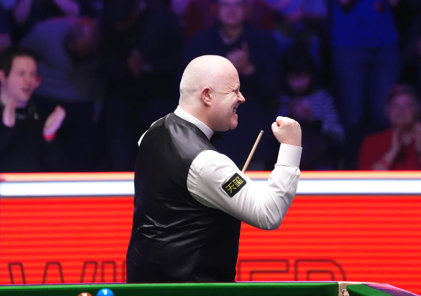 Gracz snookera w eleganckiej kamizelce świętuje zwycięstwo unosząc zaciśniętą pięść. W tle rozmyte sylwetki widzów oraz stół do gry z zielonym suknem i czerwoną tablicą świetlną.