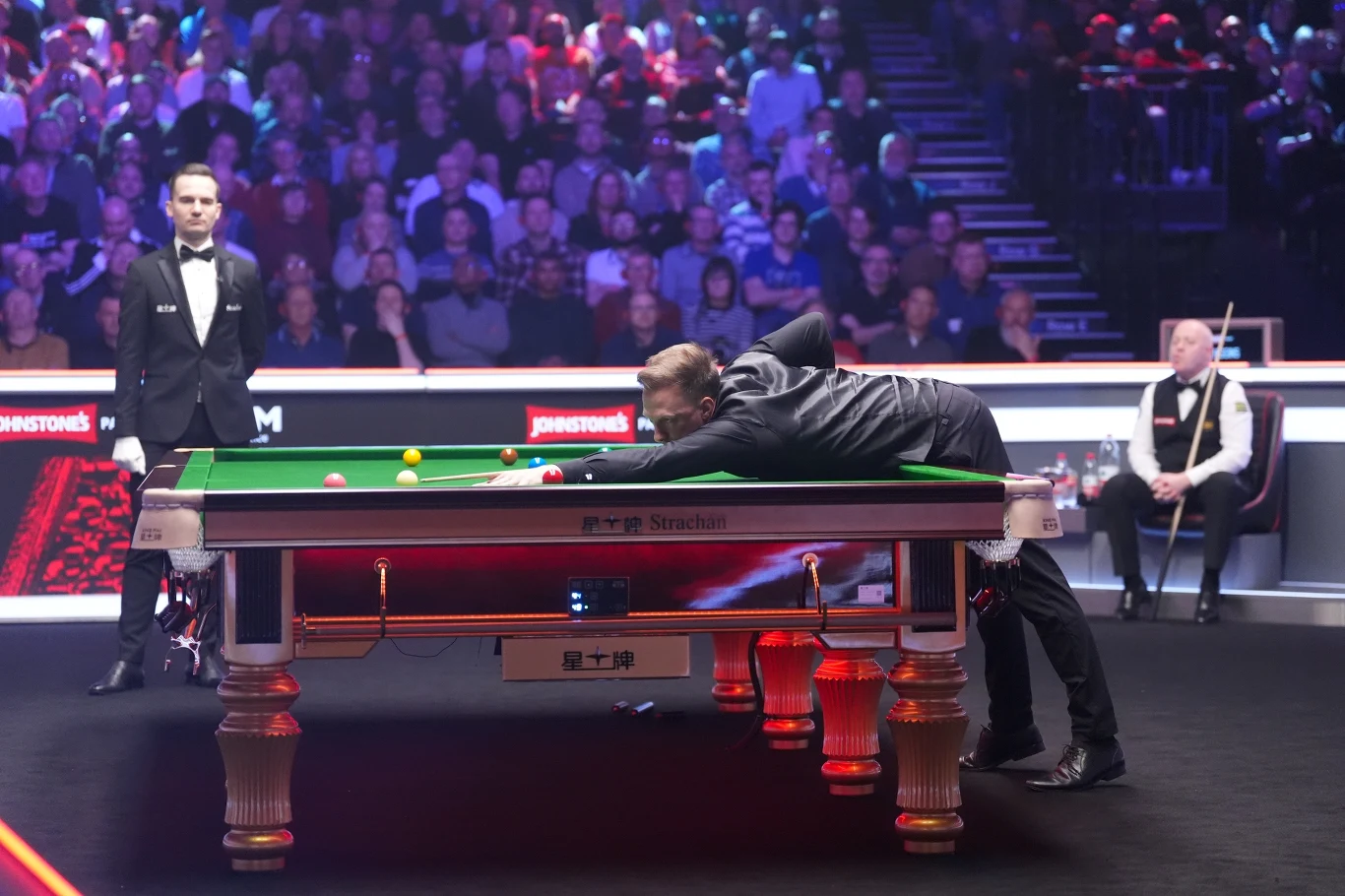 Zawodnik koncentruje się podczas wykonywania uderzenia na stole snookerowym, otoczony publicznością. Obok stołu stoi sędzia w smokingu i inny gracz siedzący na krześle w tle. Oświetlenie sceniczne podkreśla atmosferę profesjonalnego turnieju.