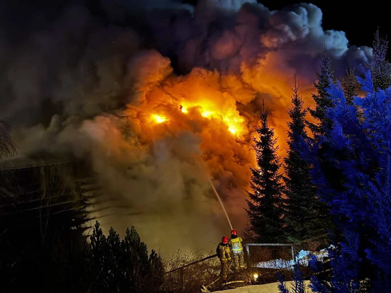 Targanice. Pożar na weselu. W lokalu wybuchł ogień. Pilna ewakuacja Intensywny pożar budynku nocą, gęsty dym oraz płomienie rozświetlające ciemność. Dwóch strażaków ubranych w mundury gaśnicze walczy z ogniem, używając węża strażackiego. W tle wysokie drzewa, część iglaków odbija niebieskie światło.