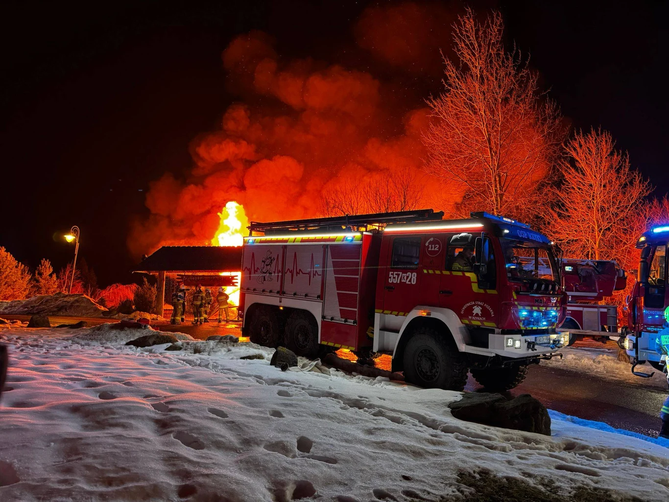 Samochody strażackie stojące nocą na zaśnieżonym terenie przed płonącym budynkiem, w tle jasne płomienie i pomarańczowe dymy, wokół kilku strażaków uczestniczących w akcji.