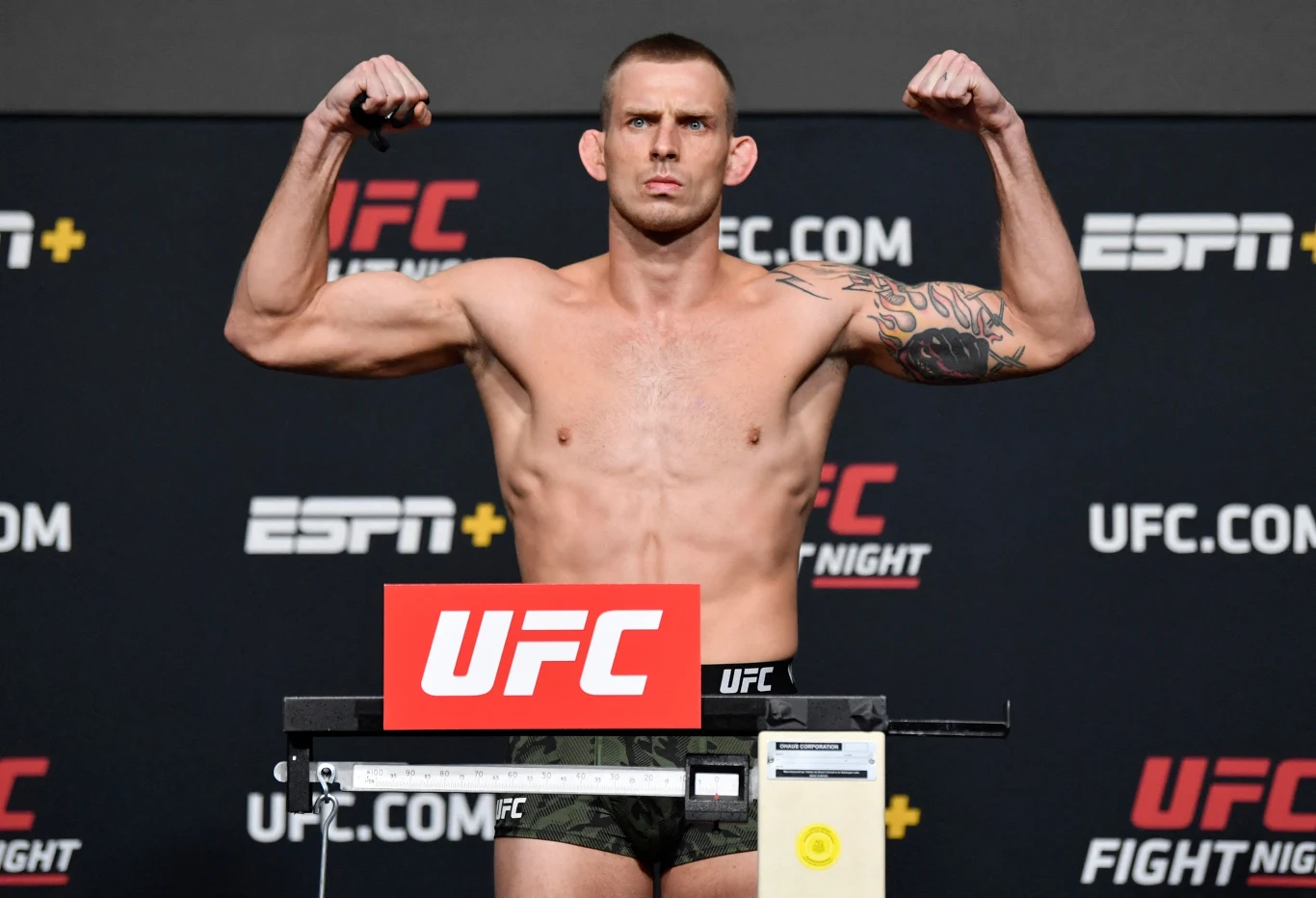 Zawodnik UFC w sportowych spodenkach stoi na wadze, prezentując muskulaturę i unosi zaciśnięte pięści w geście siły. W tle widoczne są logotypy UFC oraz ESPN.