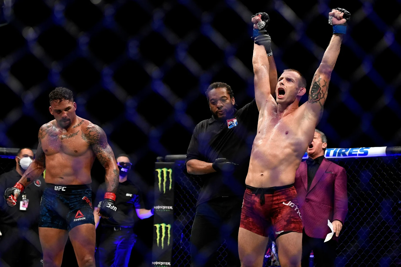 Dwóch zawodników MMA znajduje się w oktagonie, jeden z nich stoi z podniesionymi rękami w geście zwycięstwa, drugi zawodnik wygląda na zmęczonego i zawiedzionego. W tle widoczni są sędziowie i inne osoby obsługujące walkę.