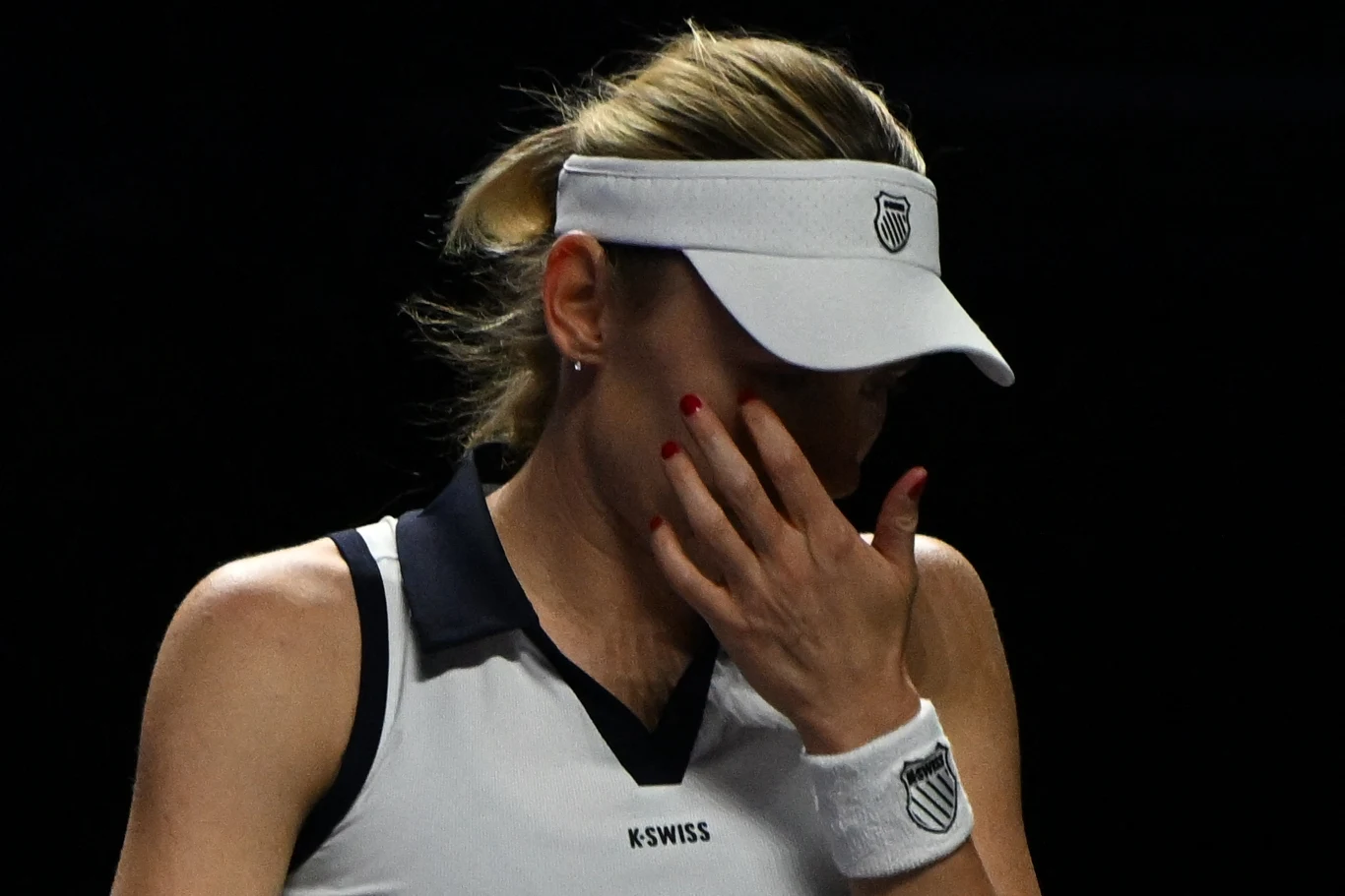 Una tenista con traje deportivo blanco y visera se cubre la cara con la mano, expresando emociones. El fondo negro centra la atención en la figura y el gesto del deportista.