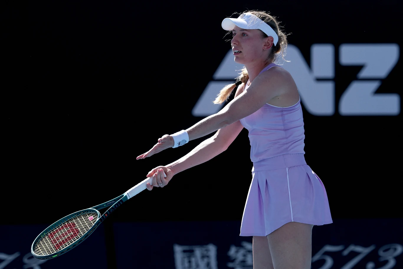 Tenisistka w fioletowym stroju sportowym z rakietą w ręku, wykonuje gest wskazujący na emocje podczas gry, w tle widoczne jest logo sponsora na ciemnym tle.