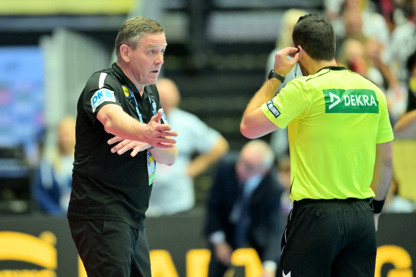 Dwóch mężczyzn w sportowych strojach prowadzi żywą rozmowę na hali sportowej, jeden z nich jest sędzią w żółtej koszulce z napisem 'DEKRA', drugi energicznie gestykuluje. W tle publiczność i rozmyte postacie.