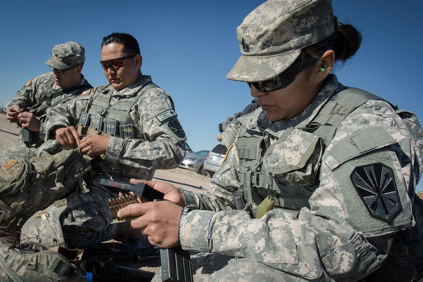 Żołnierze w mundurach polowych przygotowują amunicję na otwartym terenie, skupieni na swoich zadaniach, przy sprzyjających warunkach pogodowych.