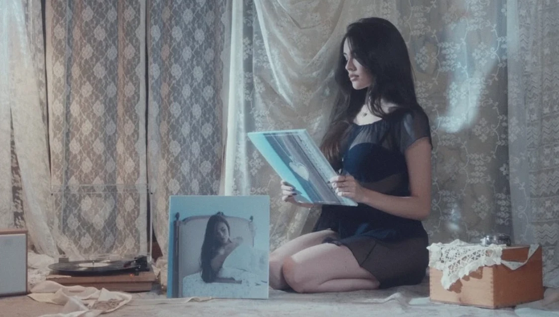 Młoda kobieta w czarnej sukience siedzi na podłodze i przegląda winylową płytę z okładką przedstawiającą jej portret, wokół niej stare pudełka oraz gramofon, całość na tle jasnych, koronkowych zasłon.