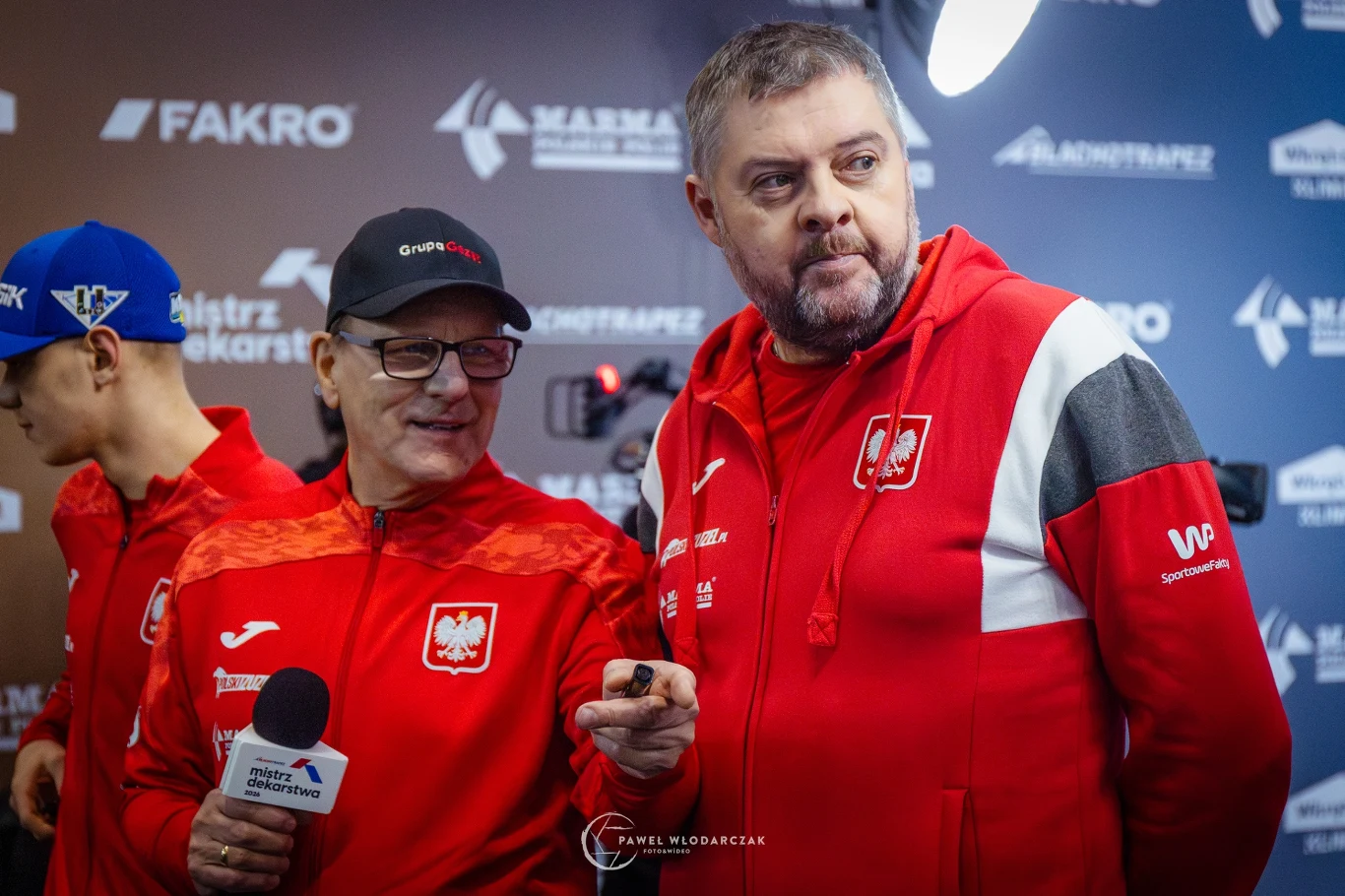 Dwóch mężczyzn ubranych w czerwone dresy z emblematem Polski w otoczeniu z elementami sponsorów i logotypami na tle mediowym, jeden trzyma mikrofon, drugi stoi obok, obaj w skupieniu.