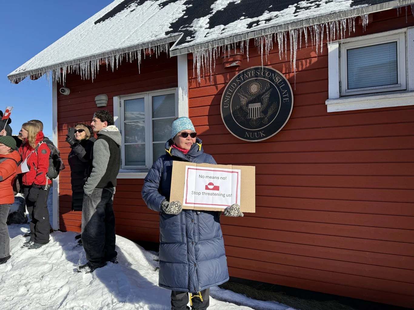 Cła na Europę rosną w tle sporu o Grenlandię, VDMA domaga się działań Brukseli (zdj. ilustracyjne) Kobieta w okularach, niebieskiej czapce i kurtce trzyma tablicę z napisem przed budynkiem z czerwoną elewacją, na którym widnieje okrągła tablica z napisem United States Consulate Nuuk. W tle kilka osób rozmawiających, dach pokryty śniegiem z soplami l...