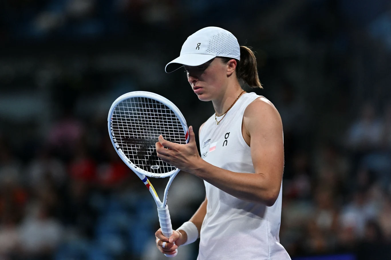 Tenisistka w białym stroju sportowym i czapce trzymająca rakietę tenisową, skupiona na korcie podczas meczu, z rozmytą widownią w tle.