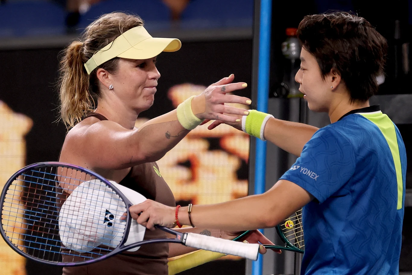 Anastazja Pawluczenkowa i Zhuoxuan Bai Dwie tenisistki obejmują się przy siatce po zakończonym meczu tenisowym, trzymając w rękach rakiety, jedna z nich ubrana jest w niebieską koszulkę, druga w brązowy strój i jasną czapkę.