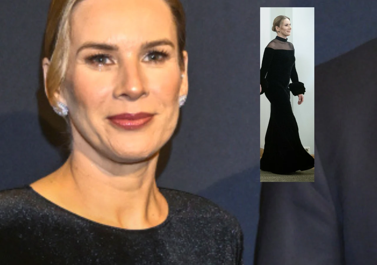 Kobieta w eleganckiej, długiej czarnej sukni z półprzezroczystym gorsetem i szerokimi rękawami, uczesane blond włosy spięte z tyłu, delikatny makijaż i wyraźnie zaznaczone usta, neutralne tło.