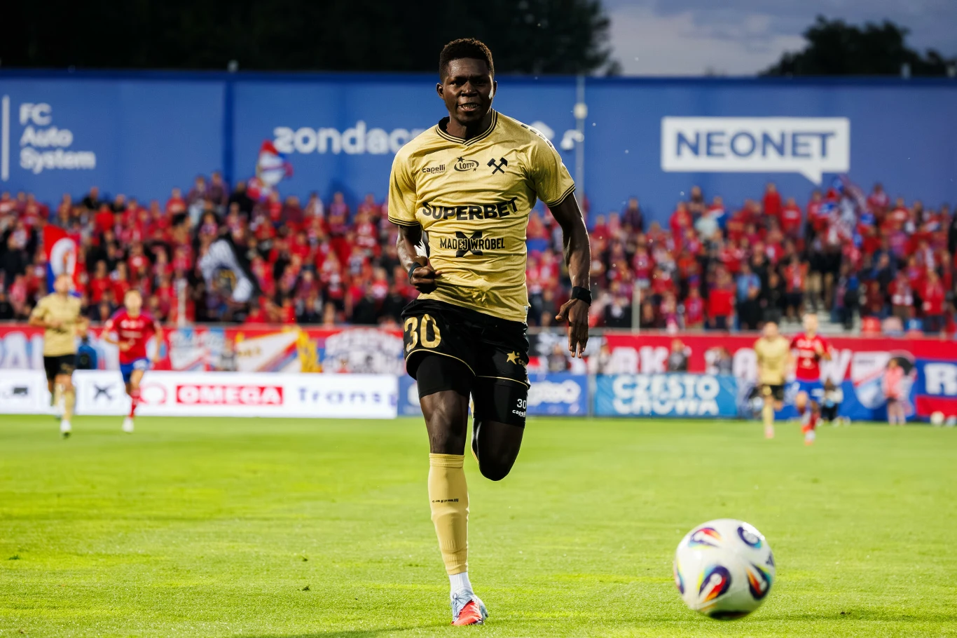 Ousmane Sow Piłkarz w złoto-czarnym stroju biegnie po murawie stadionu, skupiony na piłce toczącej się przed nim, w tle trybuny pełne kibiców w czerwonych barwach.