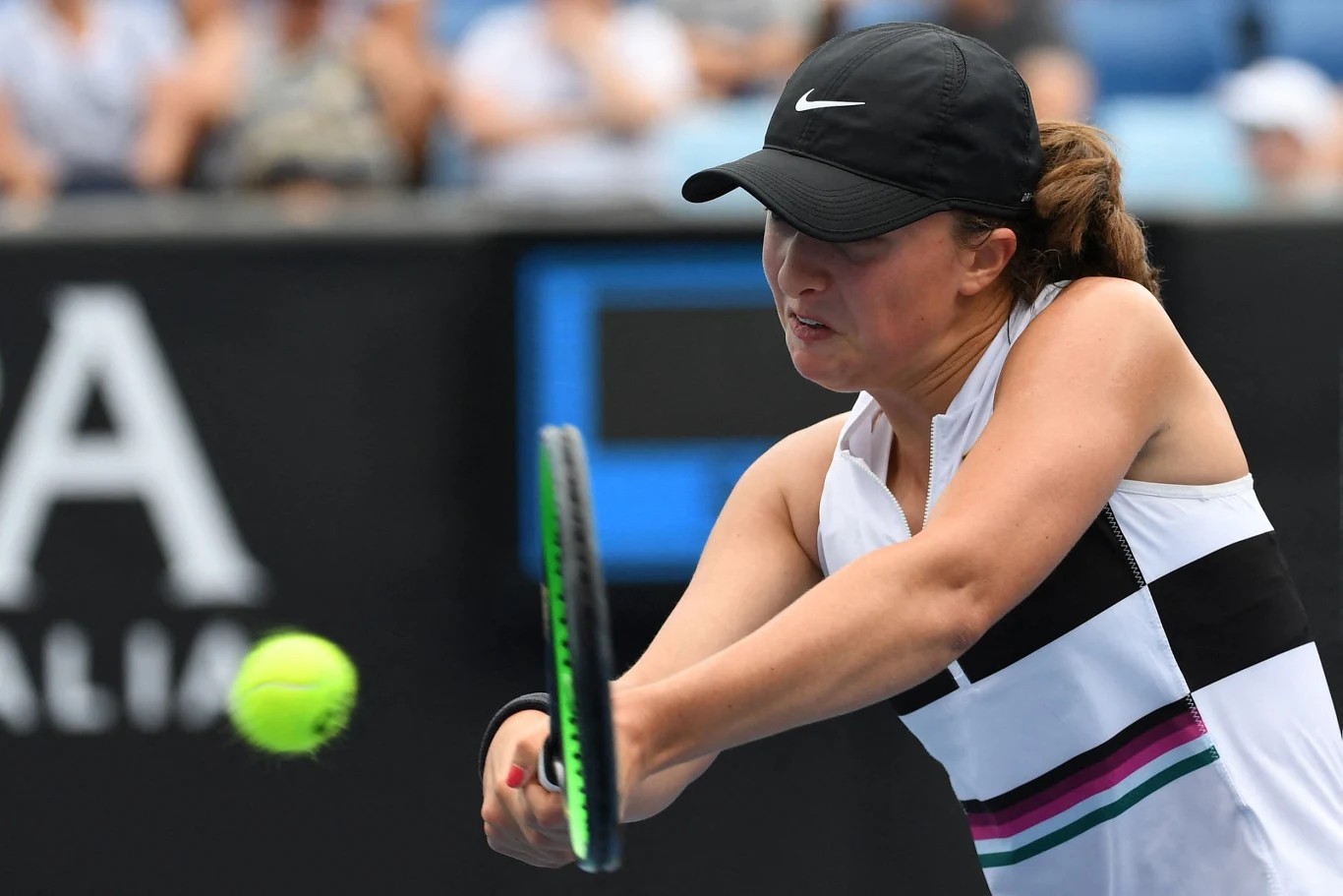 Zbliżenie na tenisistkę w trakcie dynamicznego uderzenia piłki rakietą podczas meczu na korcie tenisowym, w tle widoczna publiczność i rozmazane elementy trybun.