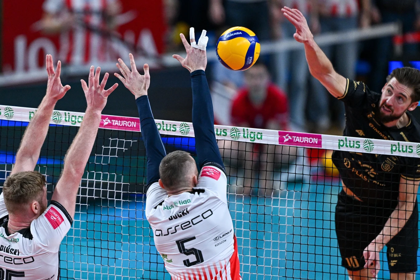 Bartosz Bednorz kontra blok Asseco Resovii Rzeszów Dynamiczna akcja podczas meczu siatkówki, zawodnik w czarnym stroju wykonuje atak, zaś dwóch graczy drużyny przeciwnej w białych koszulkach z napisem Asseco próbuje zablokować piłkę przy siatce.