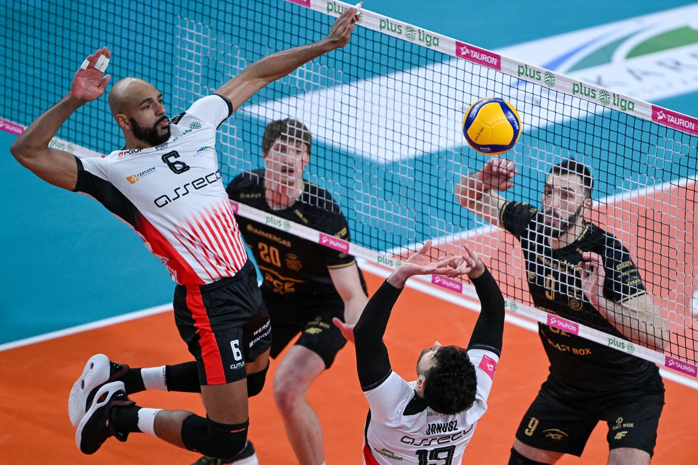 Danny Demyanenko w czasie meczu Asseco Resovia Rzeszów - PGE Projekt Warszawa Zawodnik w białej koszulce z numerem 6 wyskakuje wysoko nad siatkę podczas zaciętego meczu siatkówki, obok niego trzech przeciwników w czarnych koszulkach koncentruje się na próbie blokowania zagrania, w tle widać fragmenty boiska oraz siatkę.