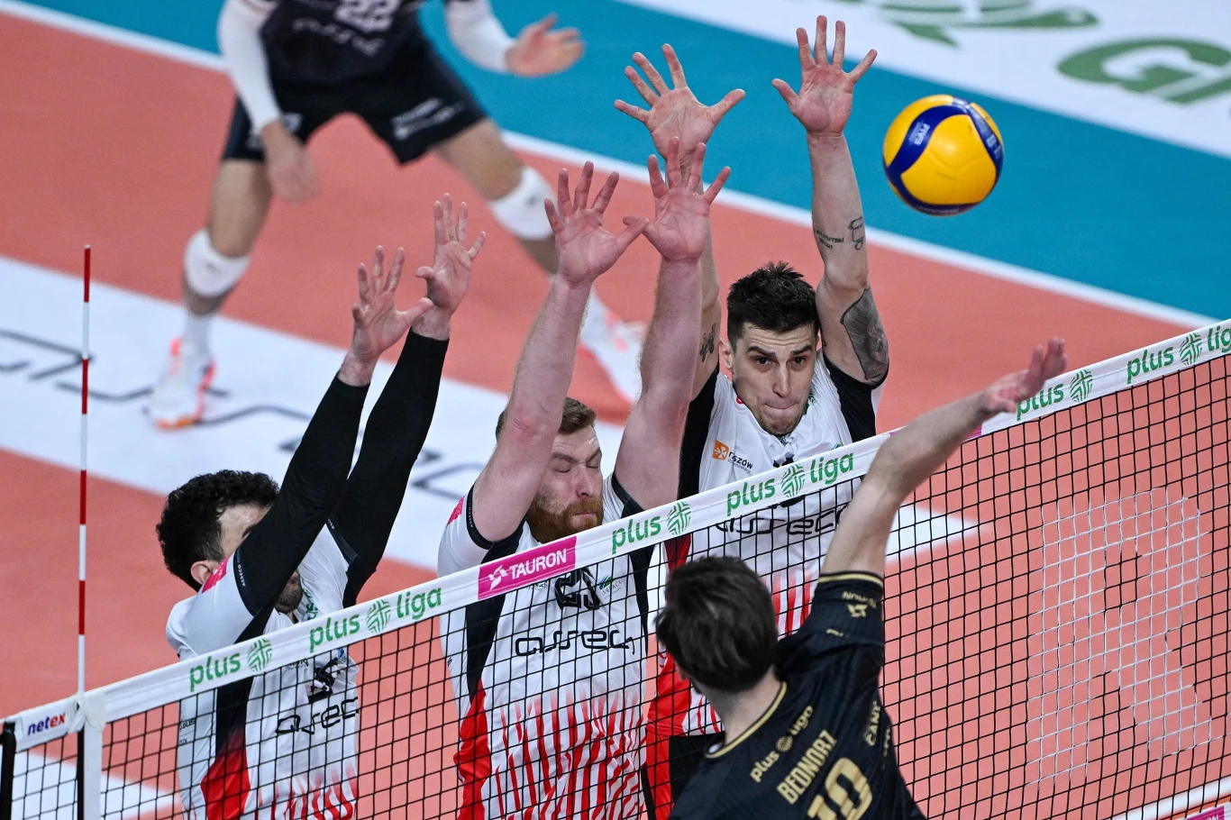 Blok siatkarzy Asseco Resovii Rzeszów Trzech siatkarzy w białych i czerwonych strojach próbuje zablokować atak zawodnika drużyny przeciwnej, piłka siatkowa znajduje się tuż nad siatką, dynamiczna akcja rozgrywa się na hali sportowej.