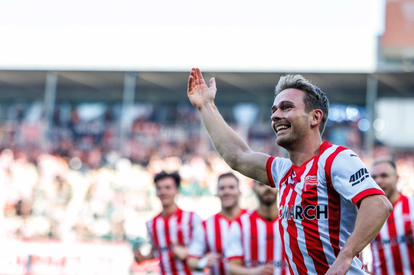 Uśmiechnięty piłkarz w czerwono-białej pasiestej koszulce, trzymający rękę uniesioną w geście triumfu, stoi na pierwszym planie przed grupą drużyny i kibicami na stadionie.