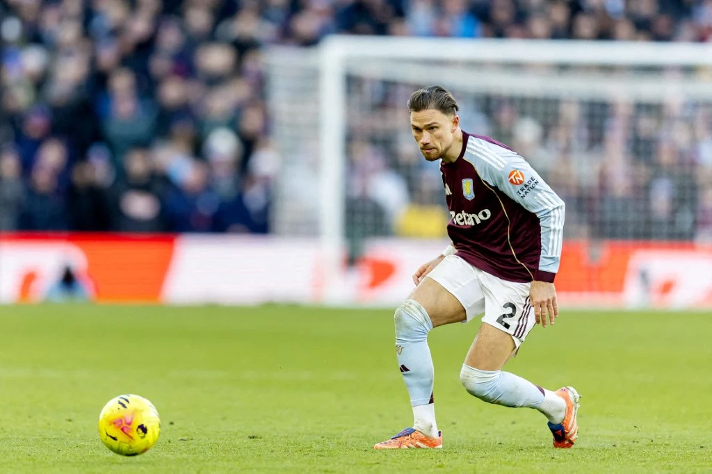 Piłkarz w bordowo-niebieskim stroju podczas meczu koncentruje się na prowadzeniu piłki na murawie stadionu, w tle widać trybuny z kibicami.