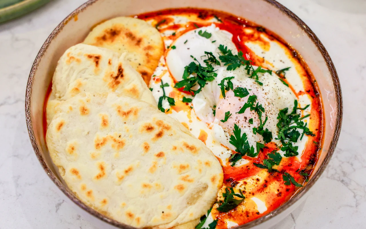 Miska z jajkami w jogurcie, z czerwonym sosem i ziołami, obok trzy chlebki naan.
