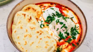 Miska z jajkami w jogurcie, z czerwonym sosem i ziołami, obok trzy chlebki naan.