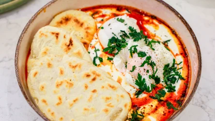 Miska z jajkami w jogurcie, z czerwonym sosem i ziołami, obok trzy chlebki naan.