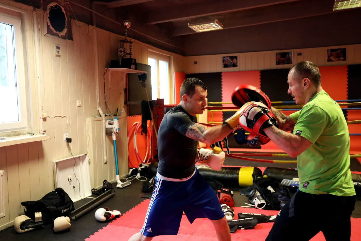 Dwóch mężczyzn trenuje boks w sali treningowej, jeden z nich uderza w tarcze trzymane przez partnera, wokół leżą sprzęty sportowe i akcesoria.