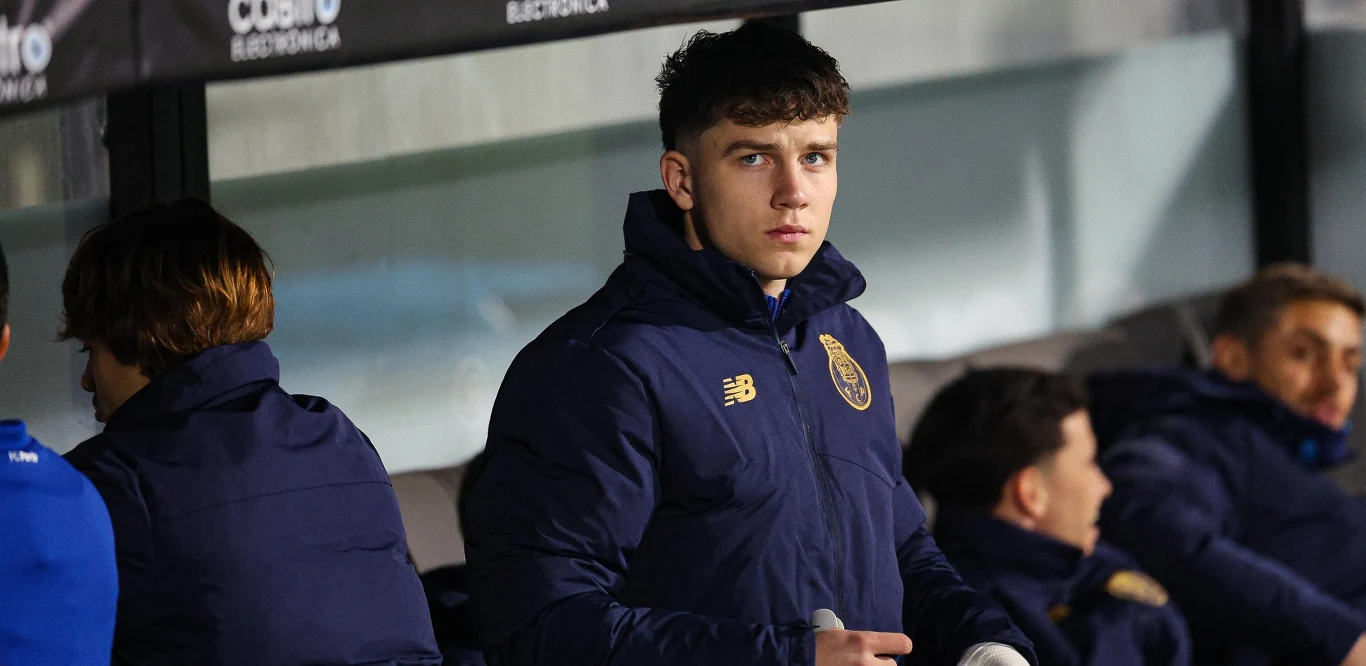 Młody mężczyzna w granatowej sportowej kurtce z herbem klubu siedzi na ławce rezerwowych, wokół niego kilku innych zawodników w podobnych strojach, tło rozmyte.