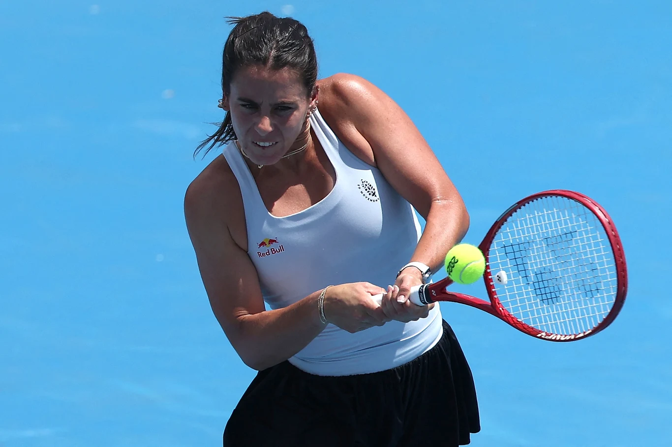 Zawodniczka w białym topie i czarnej spódniczce wykonuje uderzenie bekhendowe rakietą tenisową na niebieskim korcie, skupiona na odbiciu żółtej piłki.