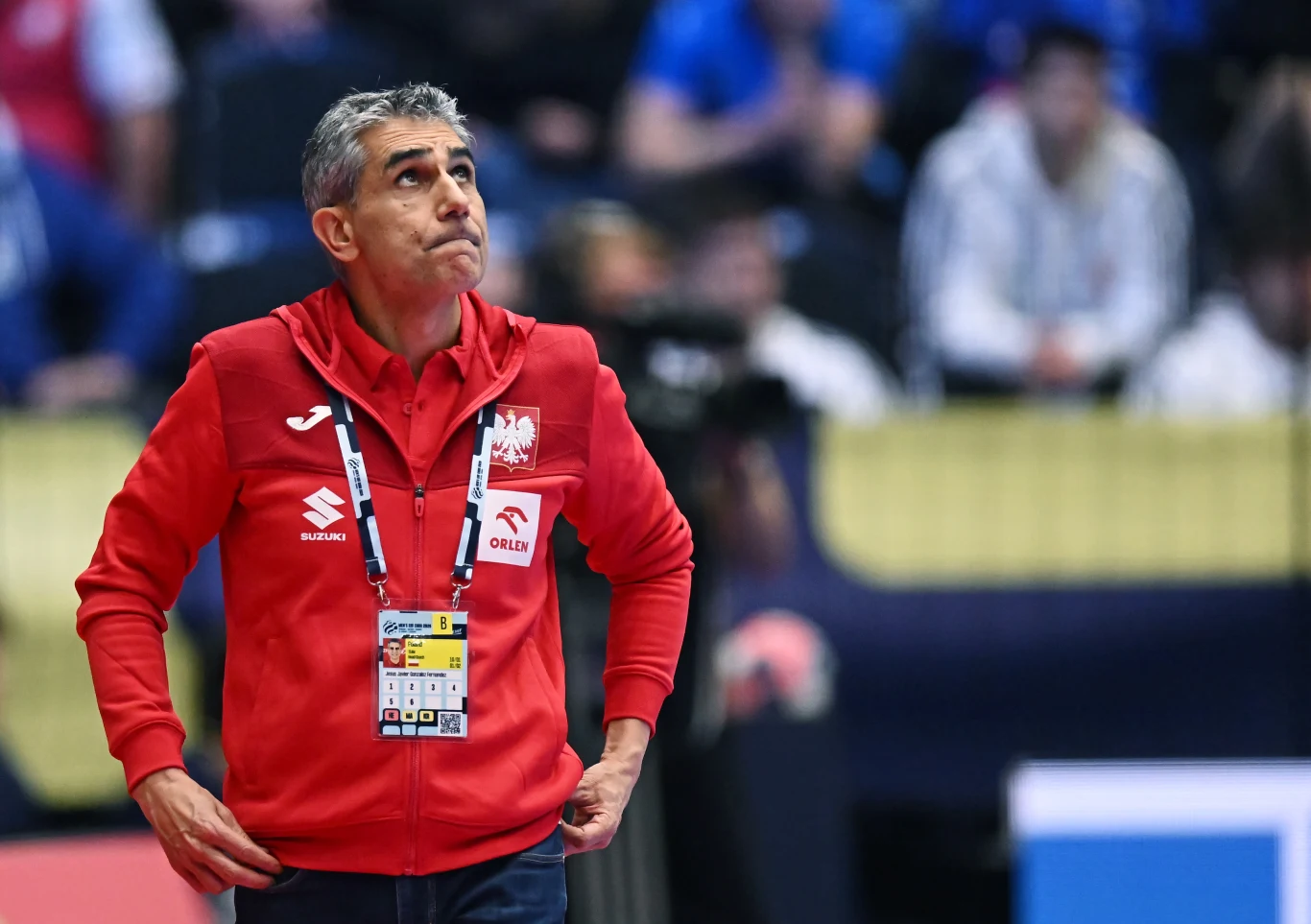 Trener w czerwonej bluzie z logiem Polskiego Związku Piłki Ręcznej stoi na hali sportowej, patrząc w górę, w tle widoczne zamazane sylwetki innych osób oraz elementy trybun.