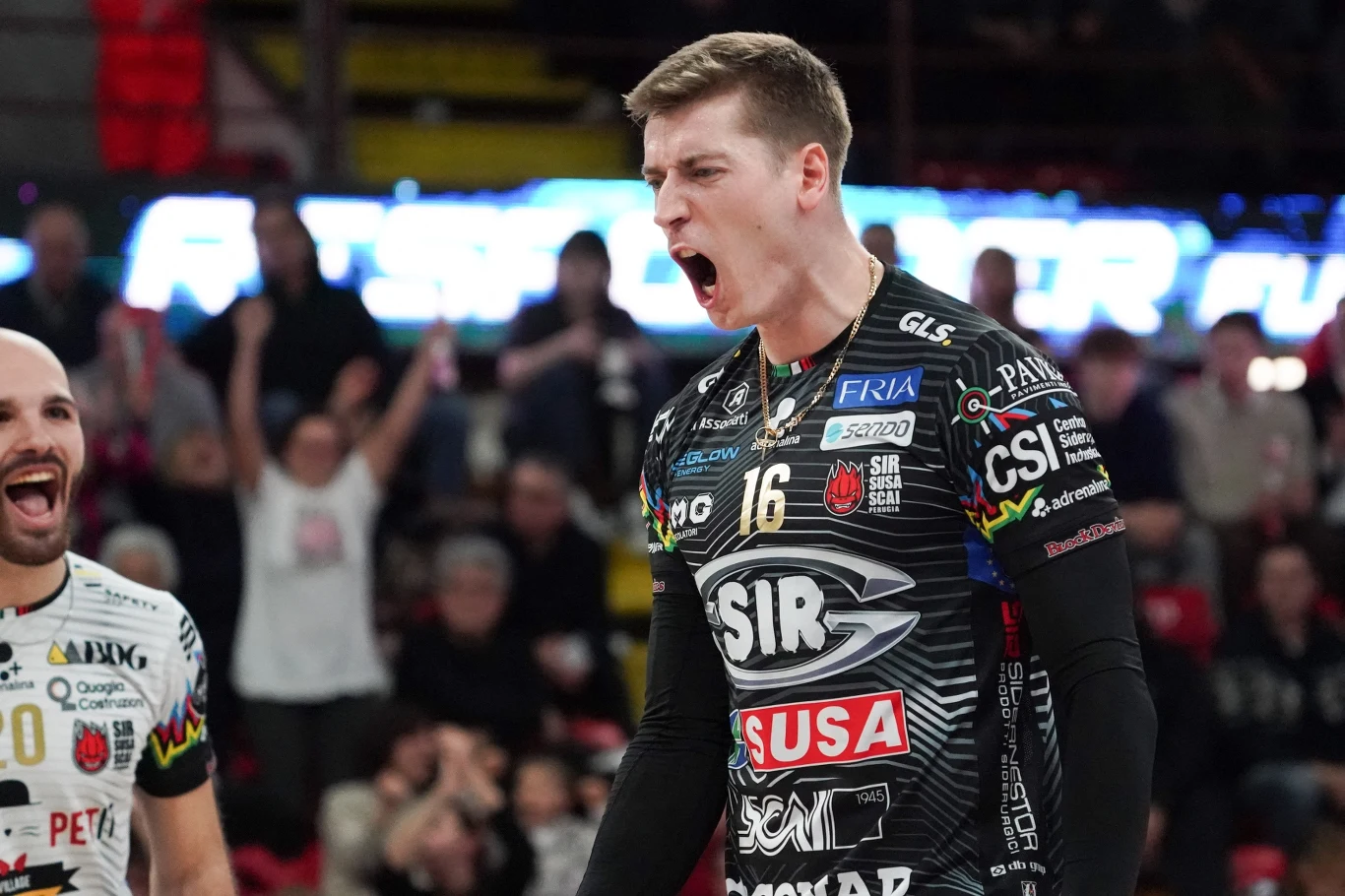 Zawodnik drużyny siatkarskiej w czarnym stroju z wyraźnym numerem 16 na piersi wyraża emocje, najprawdopodobniej po zdobytym punkcie, wokół niego widać kibiców i innych graczy na hali sportowej.