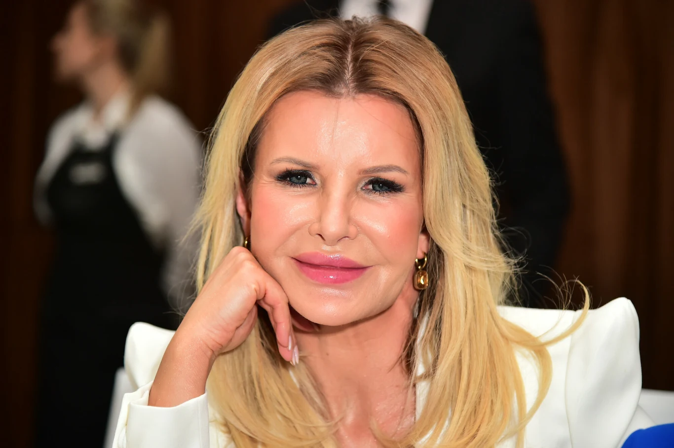 Kobieta o długich blond włosach w białej marynarce, o delikatnym makijażu i złotych kolczykach, opiera głowę na dłoni, w tle niewyraźne sylwetki innych osób.