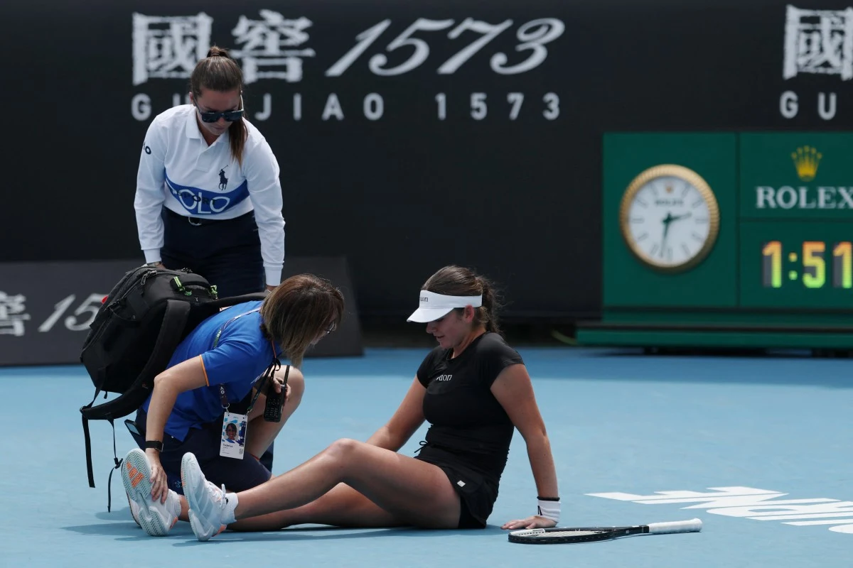 Zawodniczka tenisa siedzi na korcie, trzyma się za nogę, wokół niej są dwie osoby z personelu medycznego udzielające jej pomocy. Obok na korcie leży rakieta tenisowa, a w tle widoczne są reklamy oraz zegar pokazujący czas meczu.