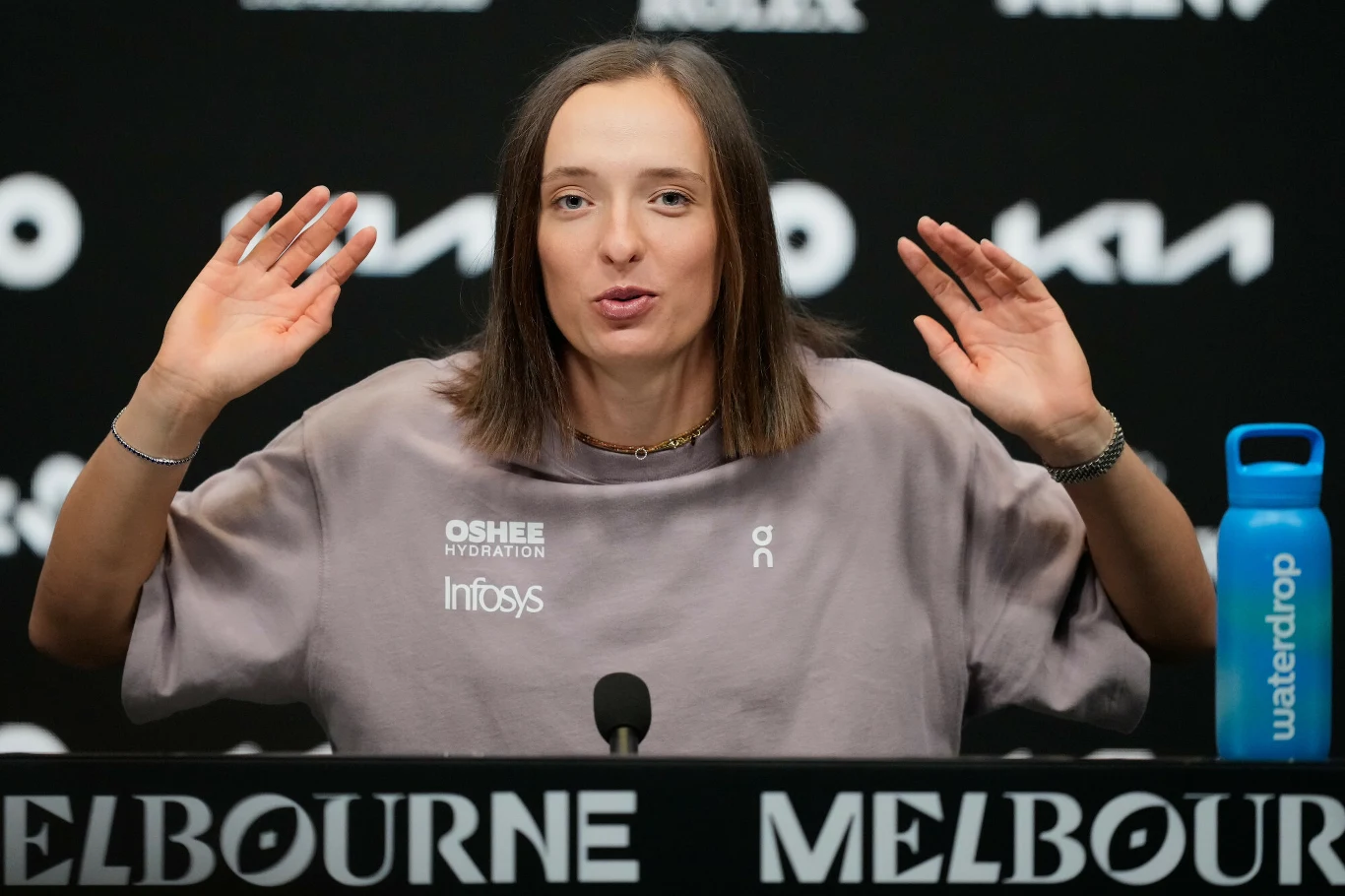 Iga Świątek Kobieta siedząca przy stole konferencyjnym z mikrofonem, unosząca ręce, w tle napisy MELBOURNE oraz logotypy sponsorów, po prawej stronie butelka z niebieską etykietą.