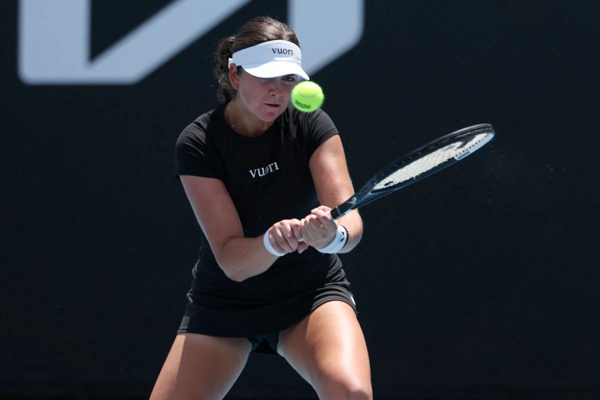 Tenisistka w czarnym stroju koncentruje się na odbiciu piłki, trzymając rakietę obiema rękoma. Piłka tenisowa znajduje się tuż przed rakietą. Na głowie opaska z logotypem. Tło ciemne, podkreślające sylwetkę zawodniczki.