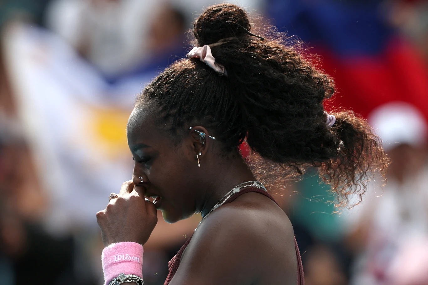 Kobieta w stroju sportowym z włosami związanymi gumką i różową opaską na nadgarstku stoi w profilu, trzymając rękę przy twarzy, tło jest rozmyte z widocznymi barwami flag.