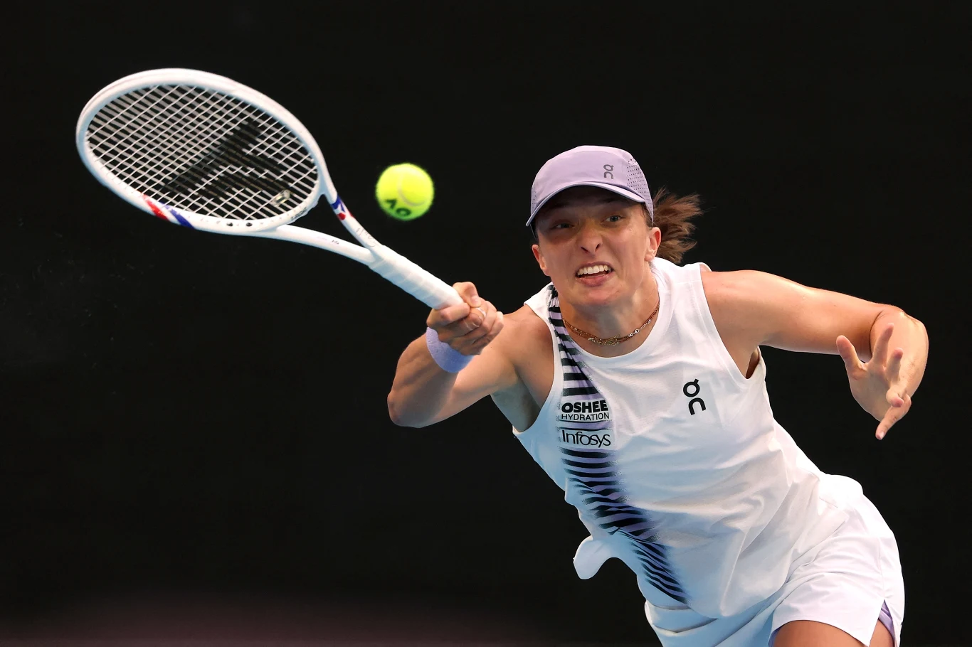 Tenisistka w białym stroju wykonuje dynamiczny ruch podczas meczu, trzymając rakietę i skupiając wzrok na zbliżającej się piłce tenisowej. Ciemne tło podkreśla ekspresję oraz sportowy charakter sceny.