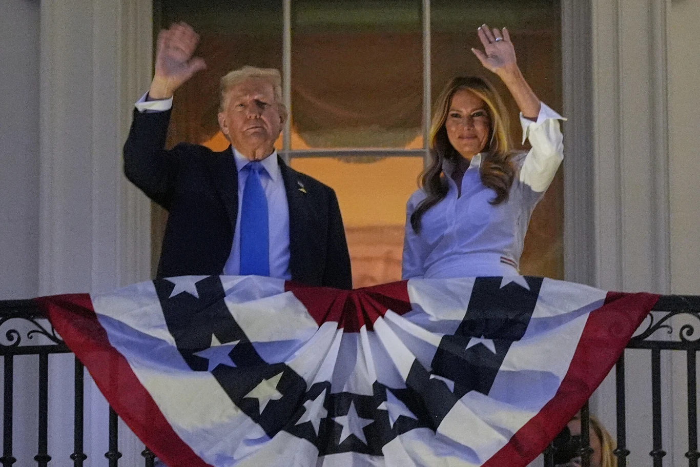 Dwie osoby stoją na balkonie udekorowanym amerykańską flagą, obie unoszą prawą rękę w geście pozdrowienia, w tle duże okno.