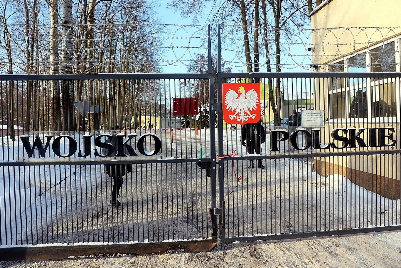 Tesle i samochody z Chin nie wjadą na tereny wojskowe w Polsce Metalowa brama jednostki wojskowej z napisem 'Wojsko Polskie' i godłem orła w koronie, otoczona drutem kolczastym. W tle zimowy krajobraz z drzewami oraz fragment budynku.