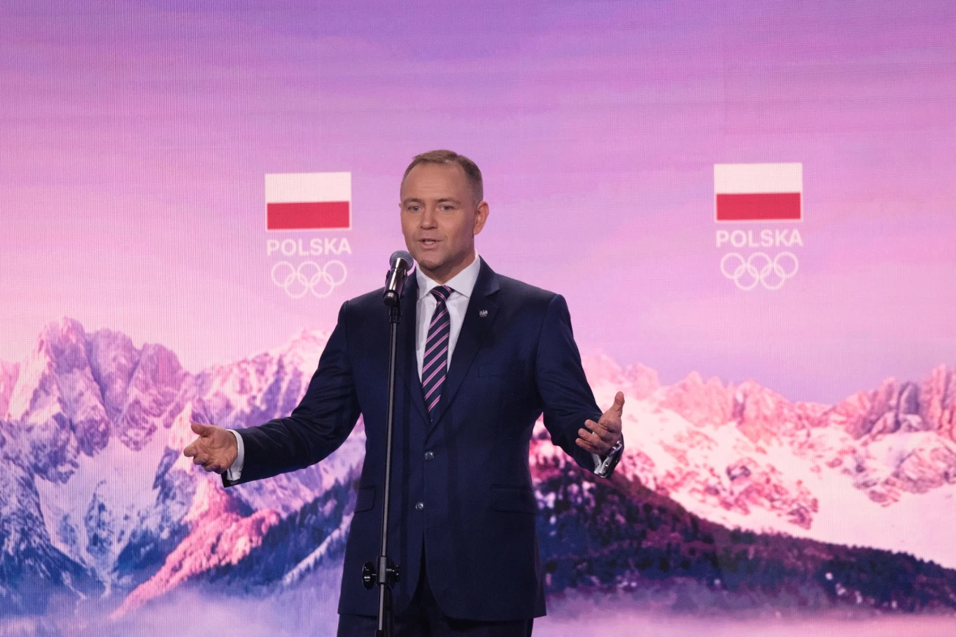 Mężczyzna w garniturze przemawia przy mikrofonie na tle gór i flag Polski oraz logotypów Polskiego Komitetu Olimpijskiego.