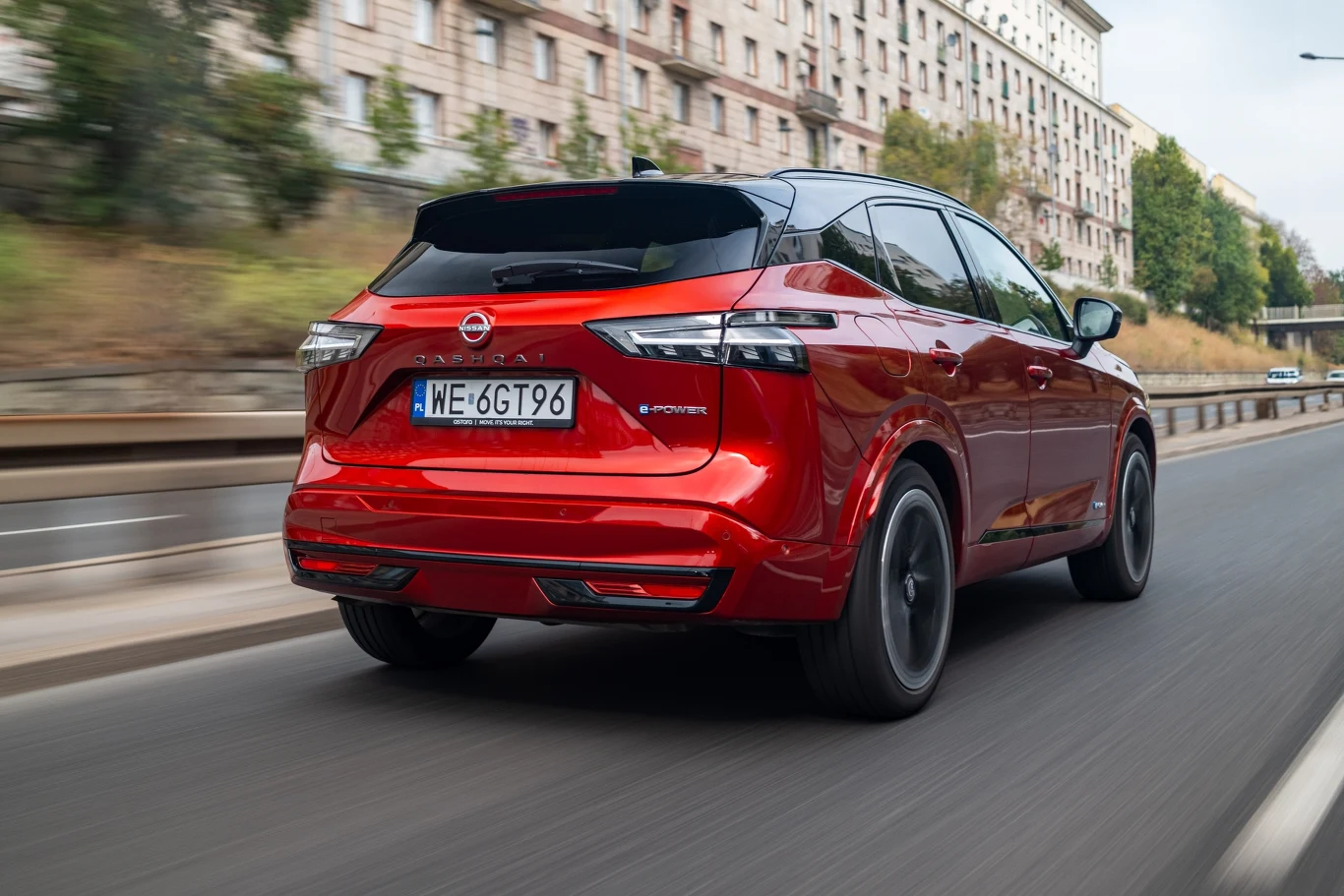 Czerwony nowoczesny samochód typu SUV marki Nissan porusza się po asfaltowej jezdni miejskiej, w tle znajdują się wielokondygnacyjne budynki mieszkalne oraz pas zieleni oddzielający ulicę.