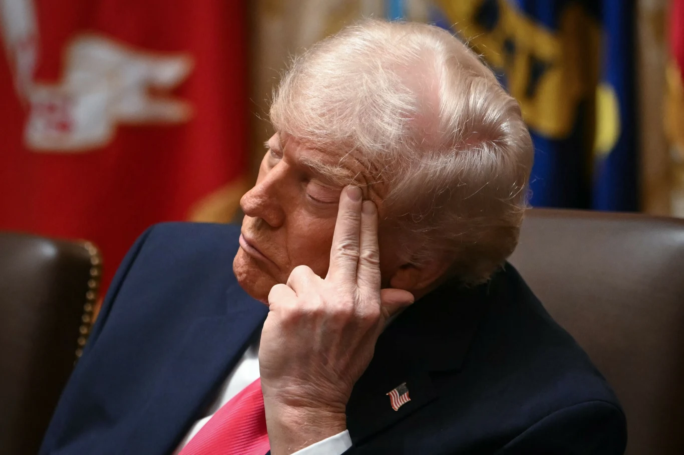 Mężczyzna w średnim wieku o jasnych włosach siedzący w eleganckim ubraniu w sali z flagami w tle, opiera głowę o dłoń z wyrazem skupienia lub głębokiej refleksji na twarzy.