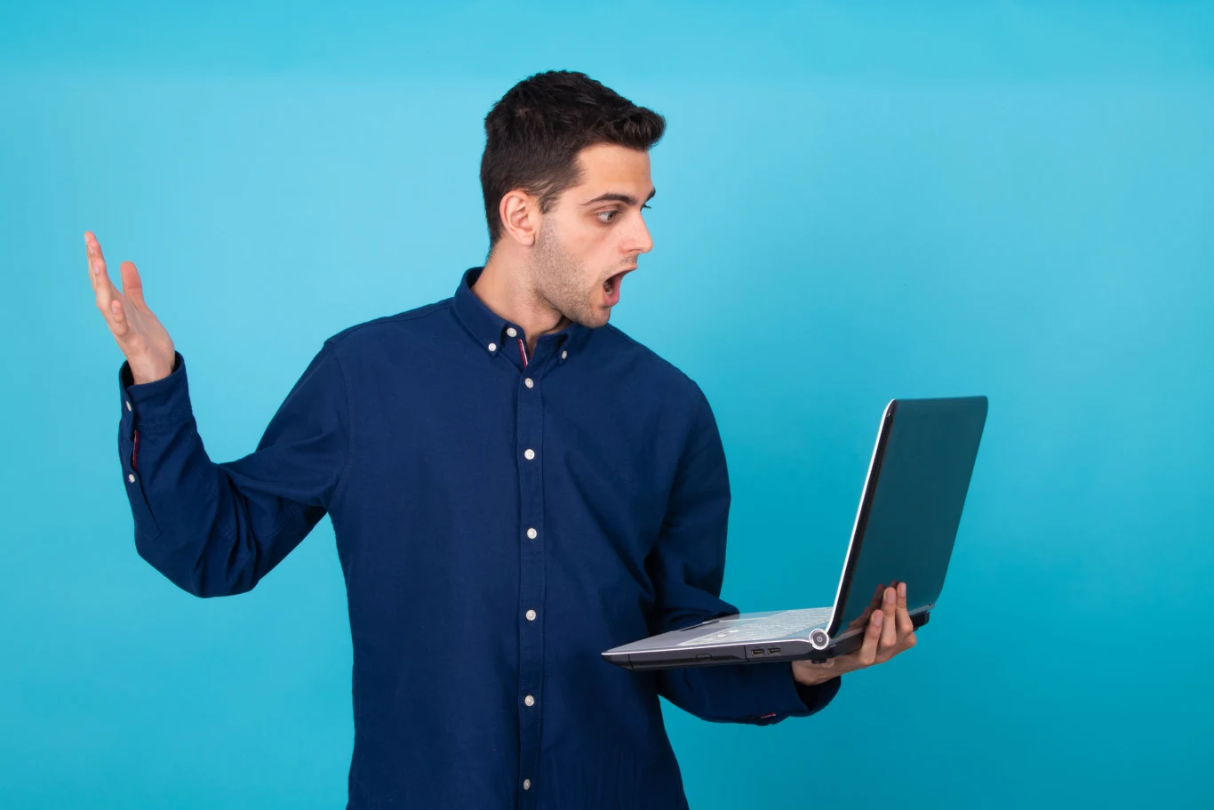 PC ciągle się zacina? Możesz precyzyjnie namierzyć podzespoły, które spowalniają cały system Młody mężczyzna w granatowej koszuli trzyma otwartego laptopa i patrzy na ekran z wyraźnym zaskoczeniem, unosząc drugą rękę w geście niedowierzania na tle niebieskiej ściany.