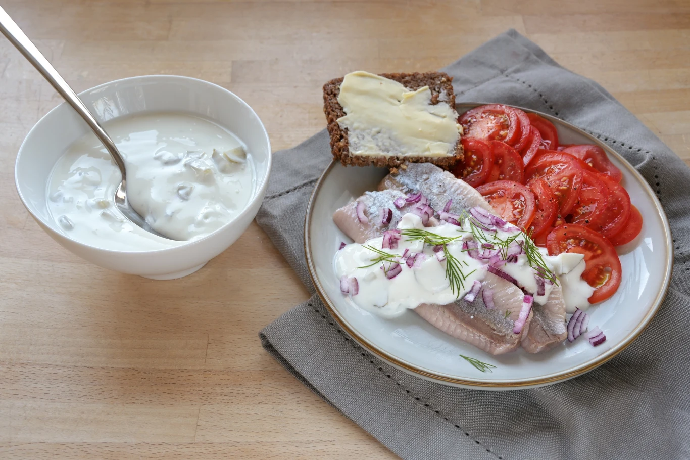 Śledzie podane z sosem śmietanowym i posiekaną czerwoną cebulą, obok plasterki pomidora oraz kromka chleba żytniego z masłem. Obok talerza miseczka z sałatką śledziową na bazie śmietany.