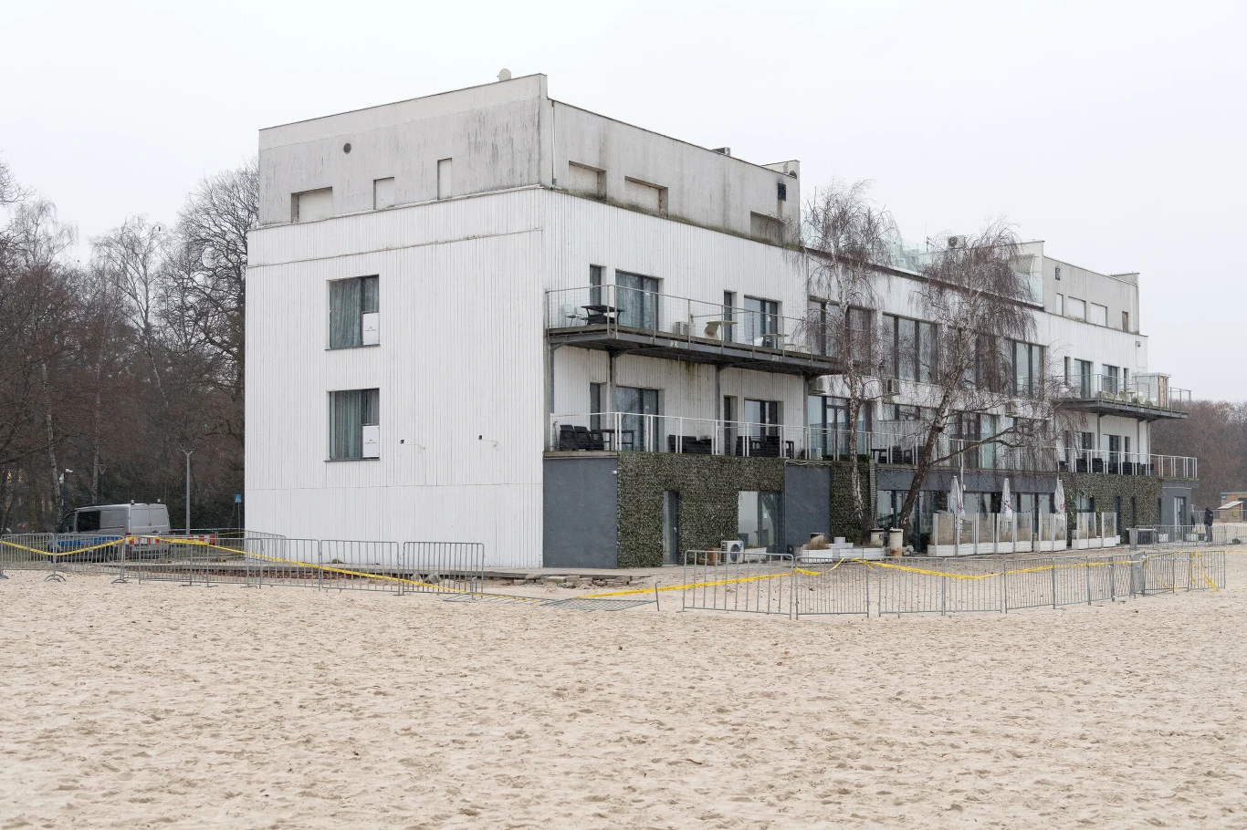 Nowoczesny, biały budynek mieszkalny z kilkoma balkonami, otoczony metalowym ogrodzeniem, ustawiony bezpośrednio przy szerokiej, piaszczystej plaży, w tle widoczne bezlistne drzewa.