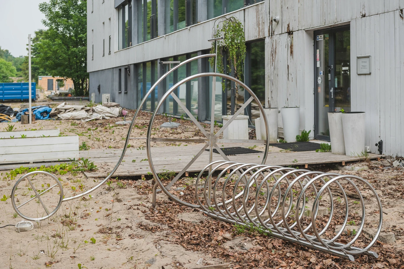 Stojak rowerowy w formie stylizowanego roweru obok nowoczesnego budynku, otoczenie zaniedbane, widoczne liście, piasek i śmieci w tle.